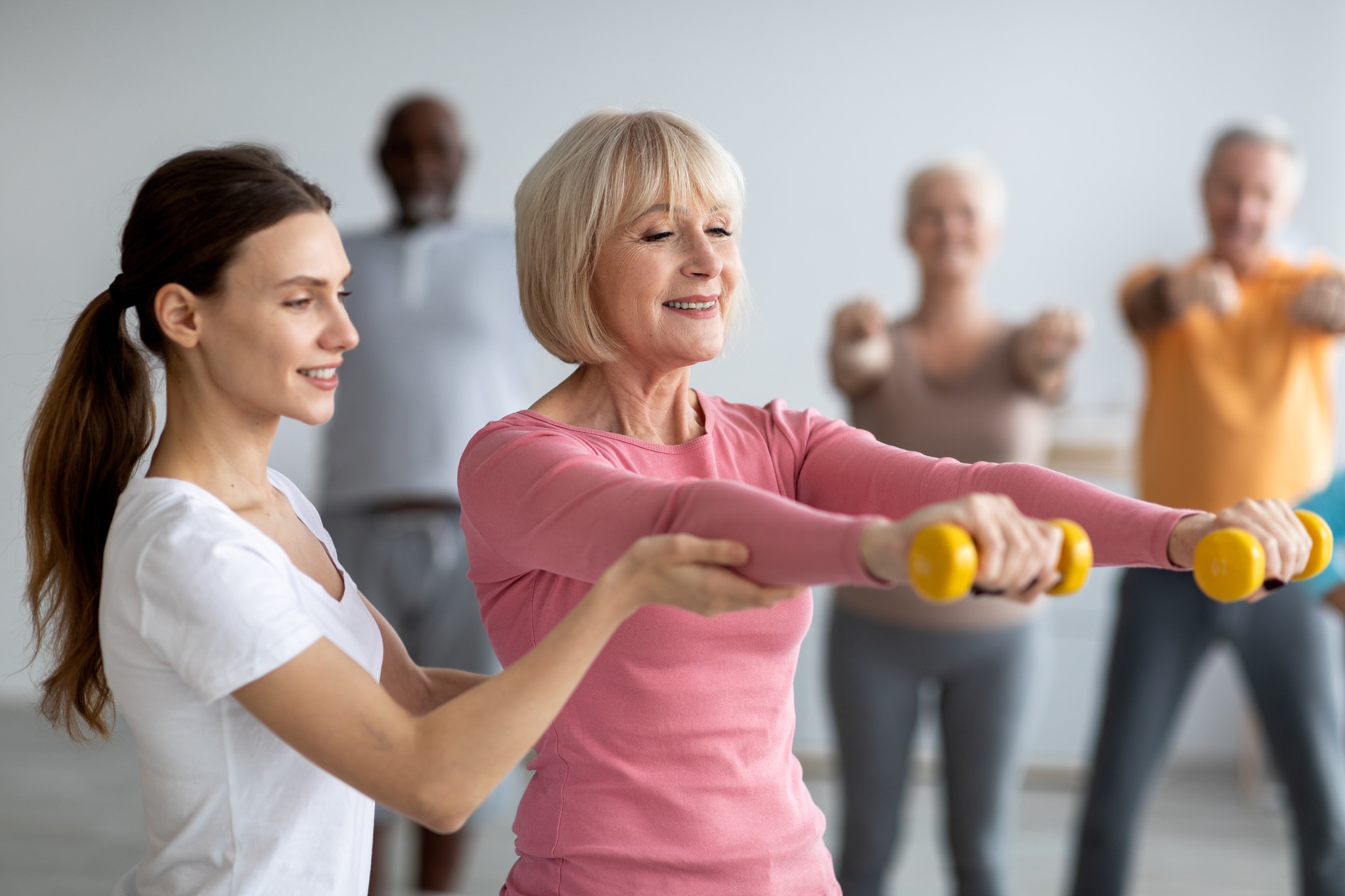 A stock image photograph of a seniors exercise class in which a lady is being helped to hold exercise weights